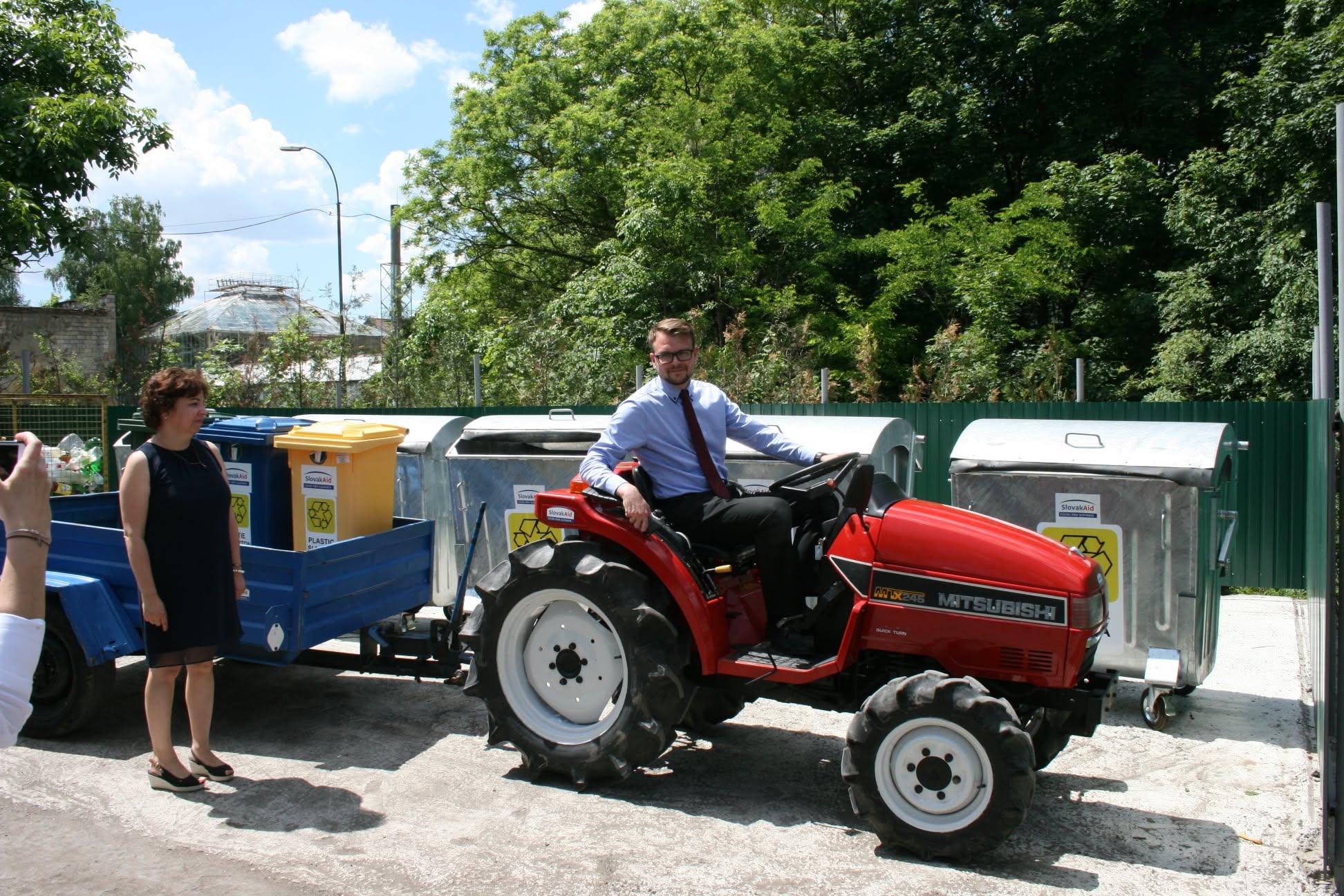 A compact tractor with a trailer next to waste-sorting containers, with people standing nearby